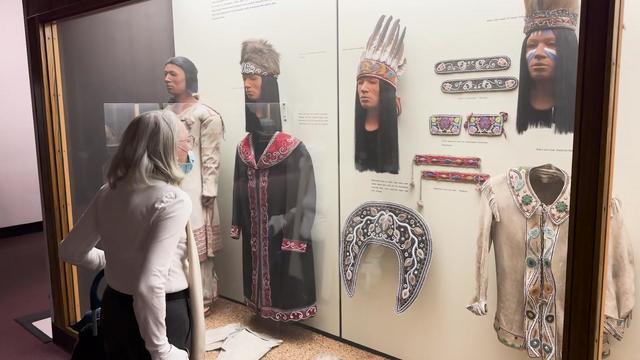 A museum visitor looks at Native American clothing items and headpieces on display behind glass. 