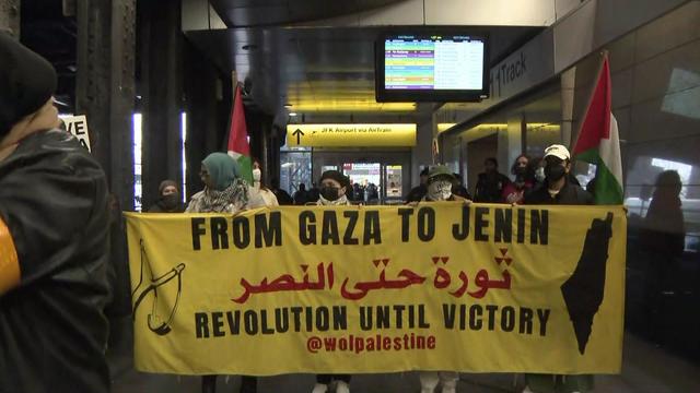 Protesters holding Palestinian flags and a banner reading "From Gaza to Jenin, revolution until victory" outside the AirTrain entrance. 