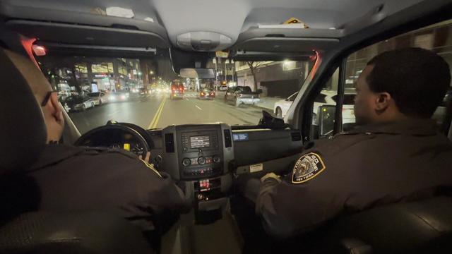 The view from the back seat of an NYPD vehicle with two NYPD officers sitting in the driver's and front passenger's seats. 