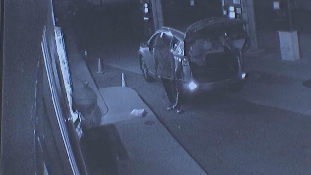 Surveillance footage shows one individual standing outside an SUV with its trunk open while another individual goes to the door of a gas station. 