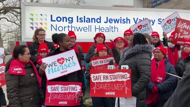 Dozens of individuals stand outside Long Island Jewish Valley Stream holding strike signs. 