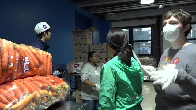 Four volunteers stand in a room containing boxes and bags of fresh vegetables. 