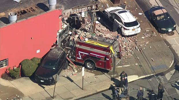 An aerial view showing a large portion of the corner of a brick building that has been destroyed. A fire truck sits partially inside the building. A white SUV sits in front of the building. 
