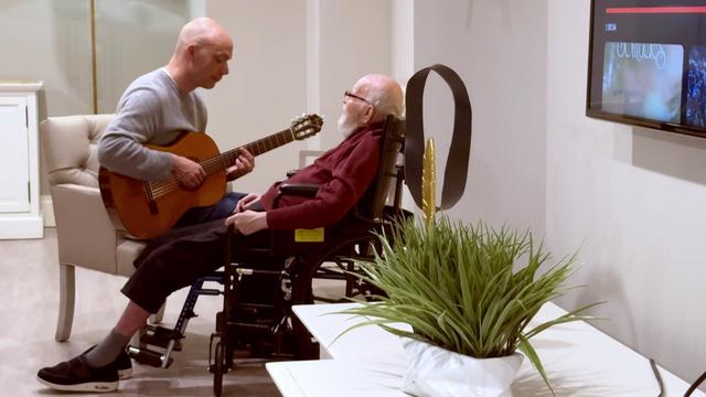 Raef Stepto sits in an armchair, playing acoustic guitar for an elderly man in a wheelchair. 