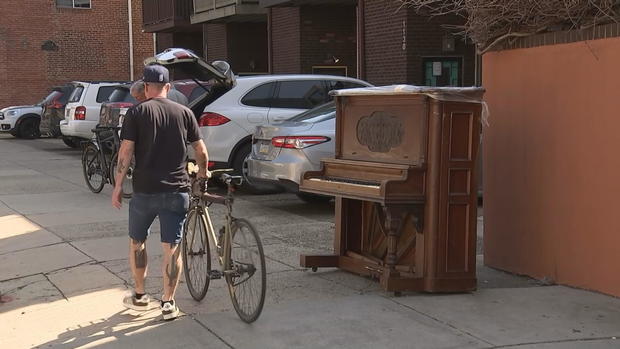 A piano sits on the street at 12th and Lombard Streets in Philadelphia. A man walking a bike passes by. 