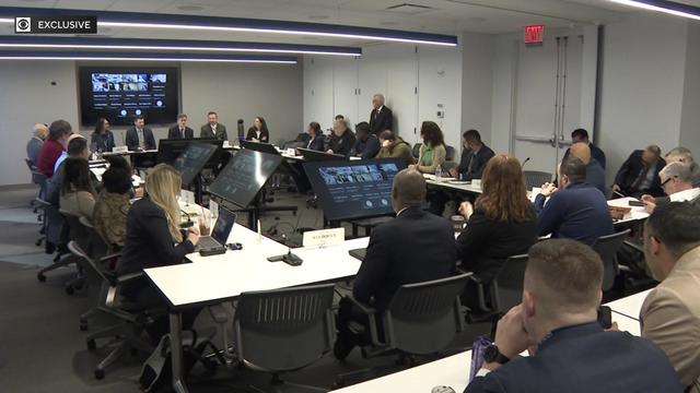 Dozens of individuals sit around a table in a meeting room. 