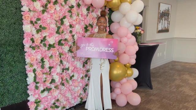 A student wearing a white and gold gown poses with a sign that says Say Yes to the Prom Dress 