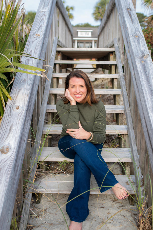 A smiling brown-haired woman sits on an outdoor staircase wearing a green sweater and jeans, her bare feet on the sand. 