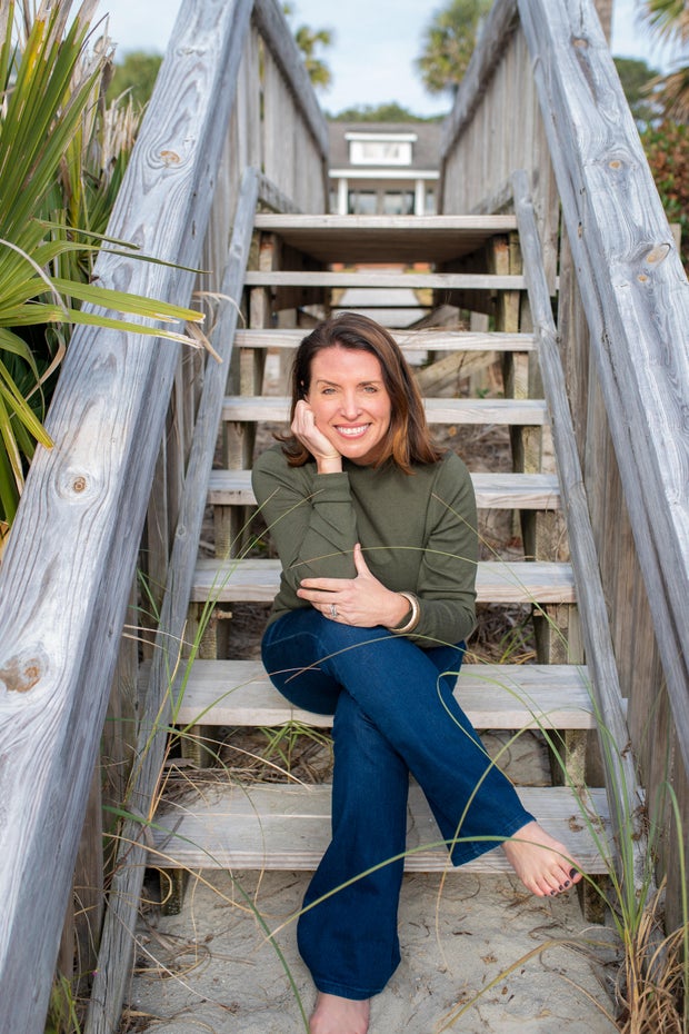 A smiling brown-haired woman sits on an outdoor staircase wearing a green sweater and jeans, her bare feet on the sand.