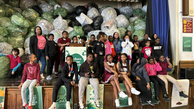 A group of students pose for a photo on a stage, in front of a pile of trash bags filled with plastic bottles. 
