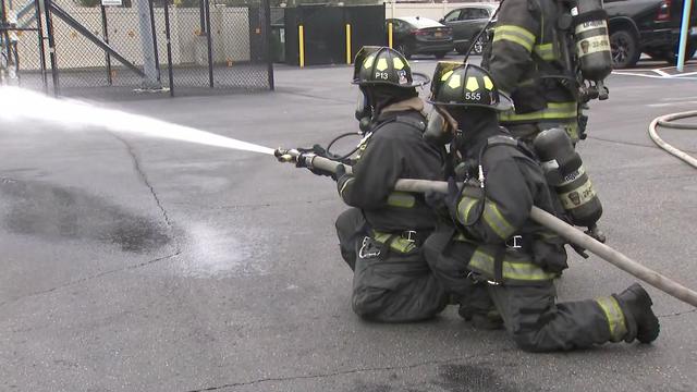 Two firefighters in full gear kneel on the ground demonstrating the use of a fire hose. 