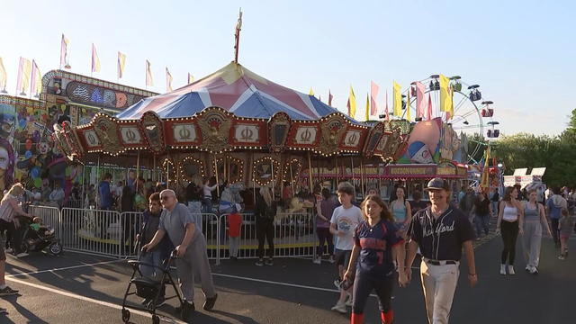 People walk around a carnival, a carousel and Ferris wheel are seen in the background 