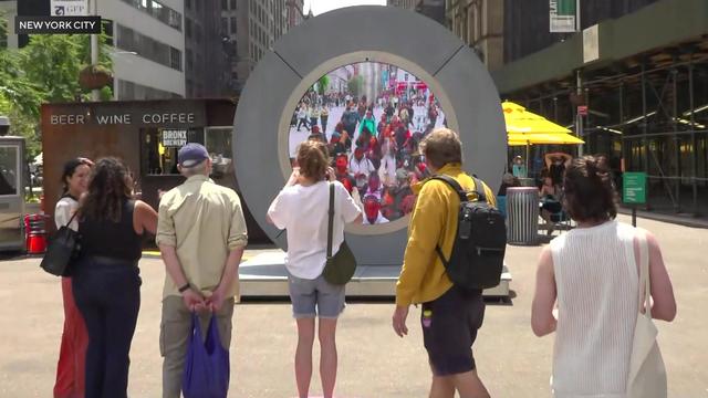 Six individuals stand in front of a large round sculpture with a video screen in the middle of it. On the video screen, dozens of people are standing in a plaza in Dublin, Ireland, seemingly looking straight at the camera. 