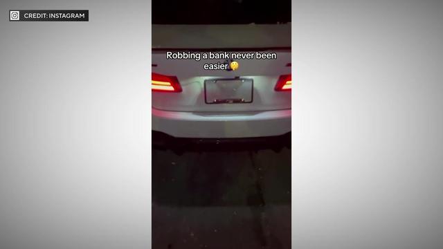 A screenshot of an Instagram video shows the back of a white BMW. A license plate cover obscures the license plate number. The caption reads "Robbing a bank never been easier" with an emoji holding a finger to its lips. 