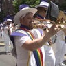 Members of a marching band wearing white uniforms with rainbow sashes perform on a Queens street. 