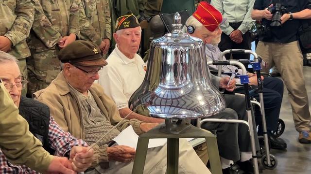 World War II veterans sit in chairs in front of a silver bell during a ceremony. 