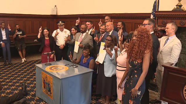 Surrounded by other people, Mayor Cherelle Parker sits at the desk where she signed the bill for a photo, everyone is holding up their index fingers to signify one 