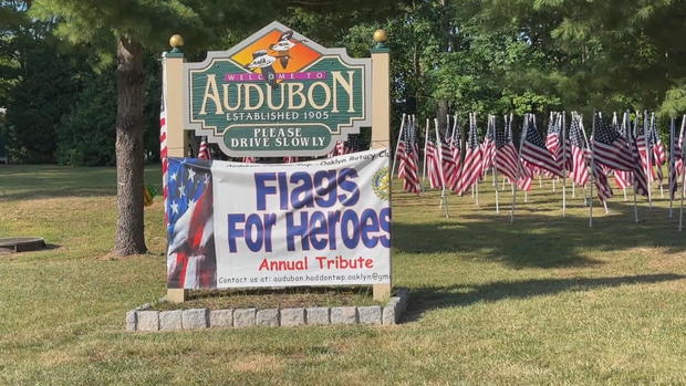 Several American flags are seen behind a sign for the borough of Audubon, which is draped with a banner that says Flags for Heroes 