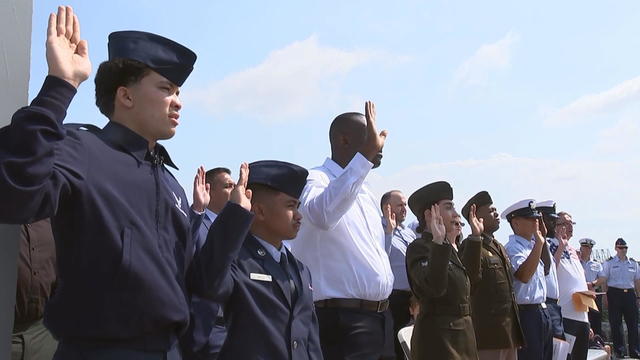 A group of people raise their right hands to take the oath of citizenship, blue skies in the background 