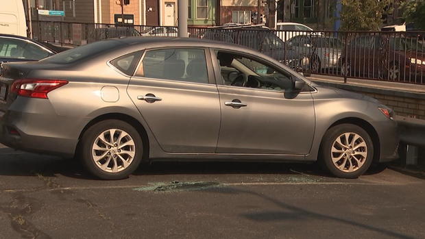 A car parked in a parking lot has a broken window on the front passenger side, and broken glass litters the ground outside the car 
