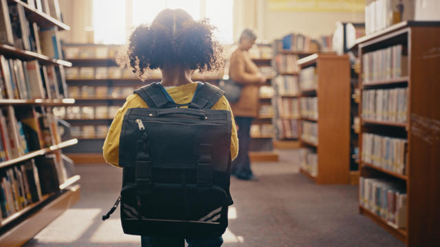 Young girl in library with backpack, books and reading for knowledge, education and learning for school. Study, kindergarten student and female kid back view, bookshelf with academic development 