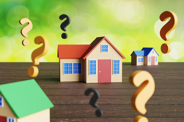Colorful miniature toy houses surrounded by yellow and black question marks on wooden table with green bokeh background. Illustration of the problems, uncertainty and unknown risks of real estates 