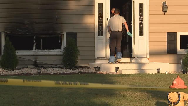 Officers enter a home that has been damaged by a fire, yellow police tape is seen in the foreground around the house 