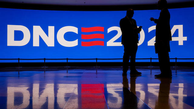 People stand in front of a sign that says DNC 2024, one person is looking at a cellphone 