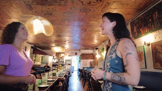 Jessi Mitchell and Sophia Litwak stand inside Symposium restaurant. Murals can be seen painted on the ceiling. 