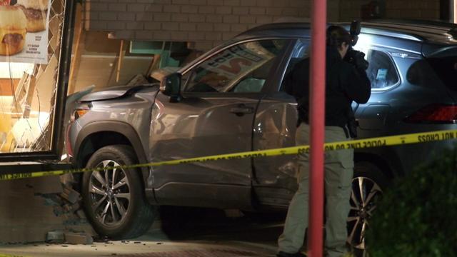 An SUV crashed into the side of a Wendy's, creating a hole in the wall. Crime scene tape blocks off the area and a police official takes a photograph of the car. 