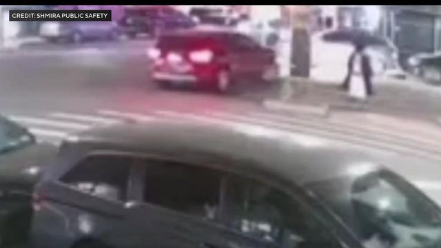 A red vehicle driving onto a sidewalk in Brooklyn as a man walks nearby. 