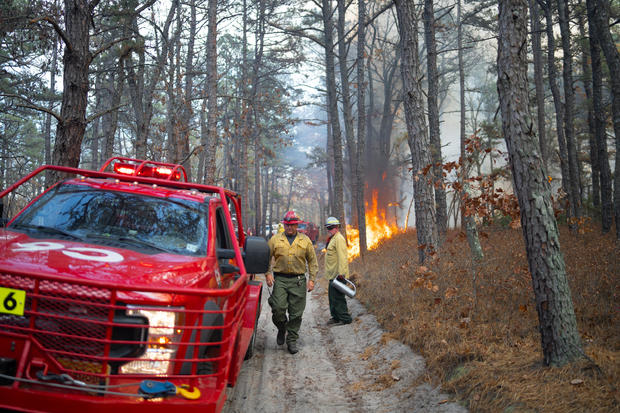 A fire truck and firefighters are seen in a forest as a fire burns in the background, hazy smoke in the background 