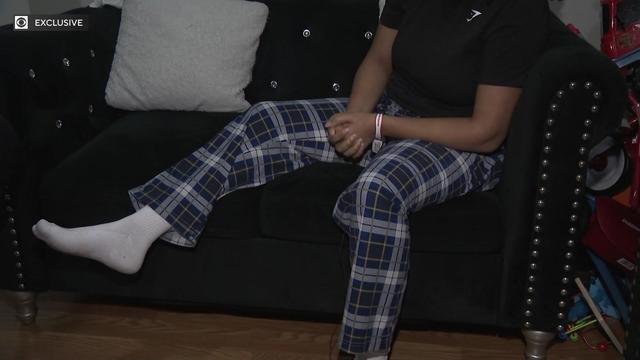 A woman wearing pajama pants, white socks and a t-shirt sits on a couch. She wears a hospital bracelet. 