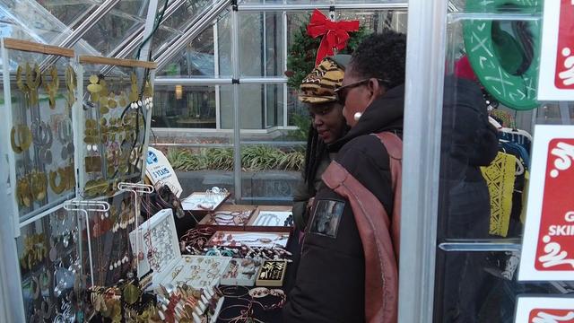 A vendor and a customer stand inside a booth at an outdoor marketplace and look at jewelry. 