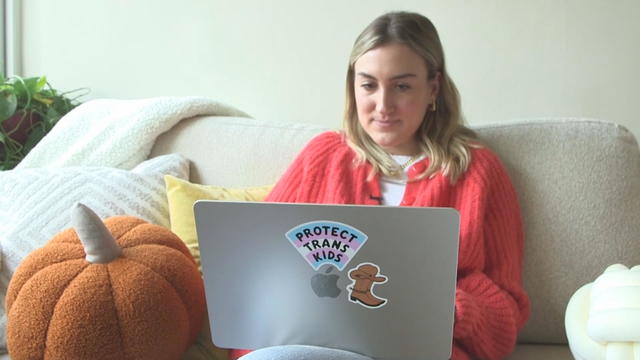 Sophia Schiaroli works on her laptop on a couch, her computer has a sticker that says Protect Trans Kids 