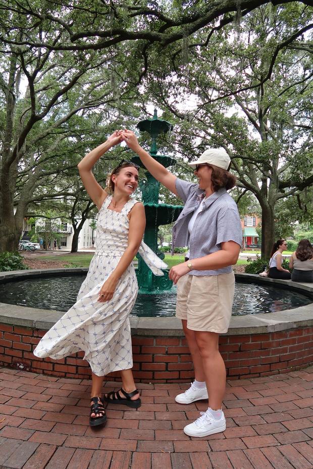 Sophia Schiaroli and Jess Plaza dance in a park in front of a fountain 