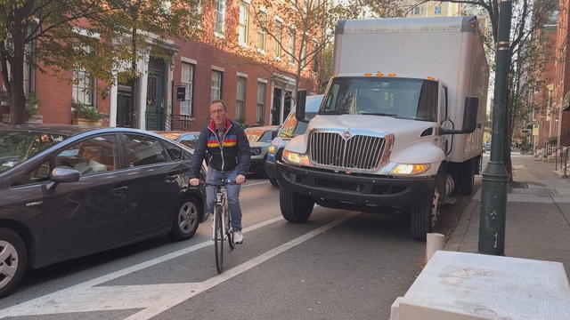 A cyclist rides between a car and a truck, which is parked in the bike lane, in Philadelphia 