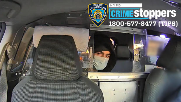 A man wearing a medical face mask sits in the back of a taxi and looks through the window into the front of the cab. 