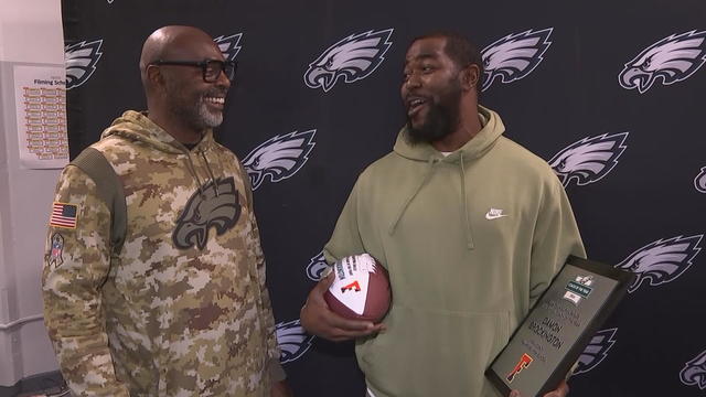 Former Eagles player Fred Barnett talks to Damon Brockington, who is holding his award, in front of an Eagles-branded backdrop 