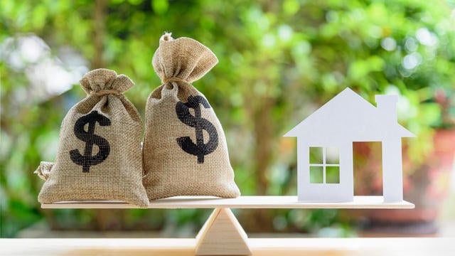 Home loan / reverse mortgage or transforming assets into cash concept : House paper model , US dollar hessian bags on a wood balance scale, depicts a homeowner or a borrower turns properties into cash 