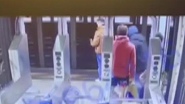 A man stands at the edge of a subway platform. A man approaches him from behind, and two bystanders stand nearby. 