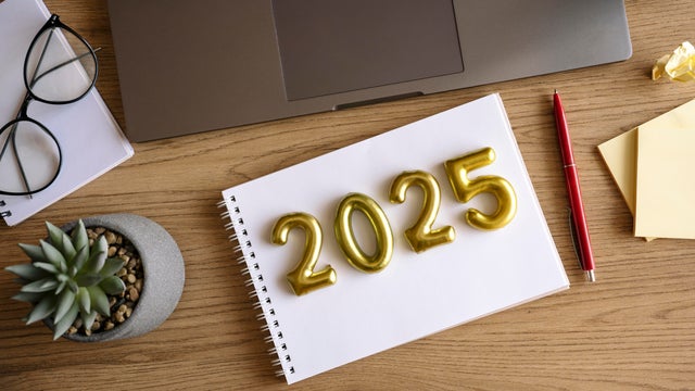 Note pad with text 2025 on wooden work desk. Planning resolutions for New Year 2025. Laptop, eyeglasses, pen. 