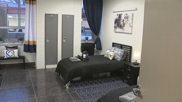 A bedroom in the wellness center, a bed with basket of toiletries and decorative pillows is seen 