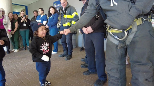 A 2-year-old boy in a FDNY beanie and FDNY sweatshirt fist bumps a first responder. The boy has a large bandage covering his right hand. 