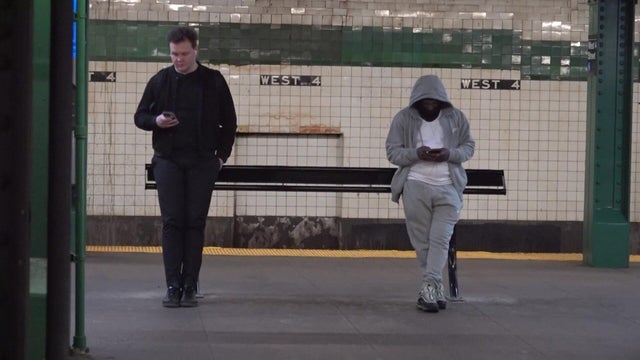 Two individuals lean against a slightly angled black metal "leaning bench" on a subway platform. 