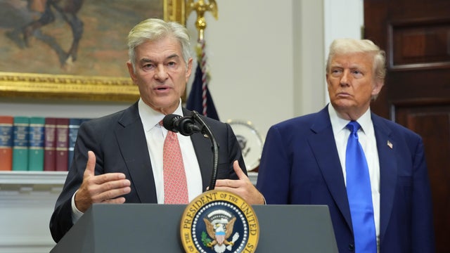 Dr. Mehmet Oz speaks at a presidential podium as President Trump looks on during an event in the Roosevelt Room at the White House 