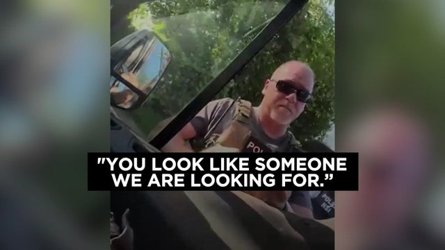 An ICE agent stands at the open passenger side window of a vehicle. A caption reads, "You look like someone we are looking for." 