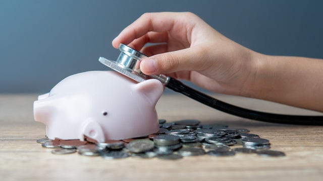 A hand holding a stethoscope listening to a broken piggy bank lying on a pile of coins. 