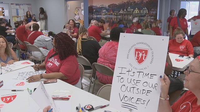People wearing red t-shirts work on posters, one holds up a sign with the Philadelphia federation of teachers logo that says It's time to use our outside voices 