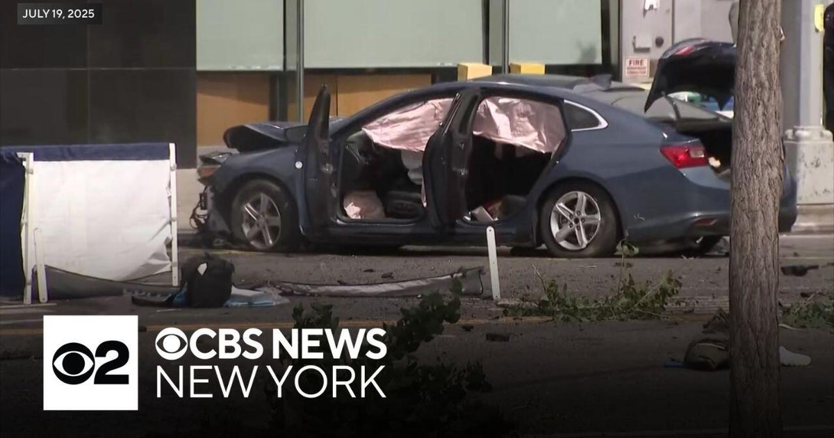 Blue four-door sedan parked on a city street with all passenger doors open, side airbags deployed and the trunk open. Debris and leafy branches are scattered on the pavement in front of the vehicle; a white privacy screen stands to the left. A tree trunk and building facade are visible at right and background.